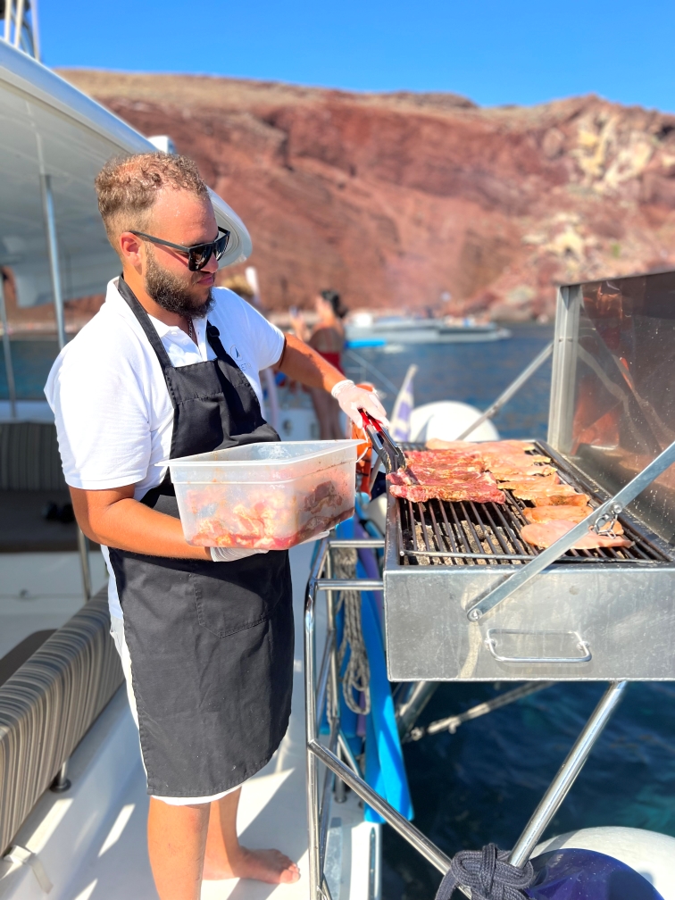 Crew member grilling meat while sailing near red beach in Oia